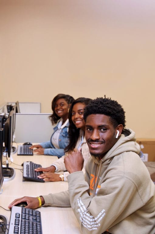 three young black students smiling at the camera while working in a computer lab