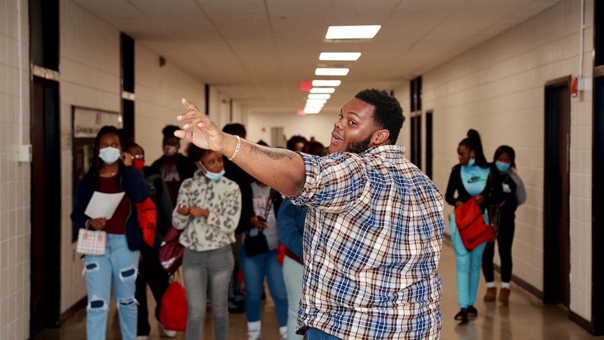 A young, black male college student guiding high school students down a hall while walking backwards