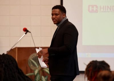 A young black male college student wearing a thick turtle neck addresses a group of high school students