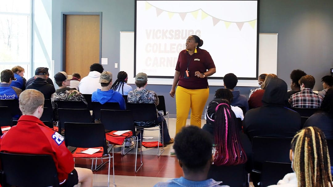 A black female college administrator, wearing a maroon shirt and gold pants, addresses a group of seated high school students