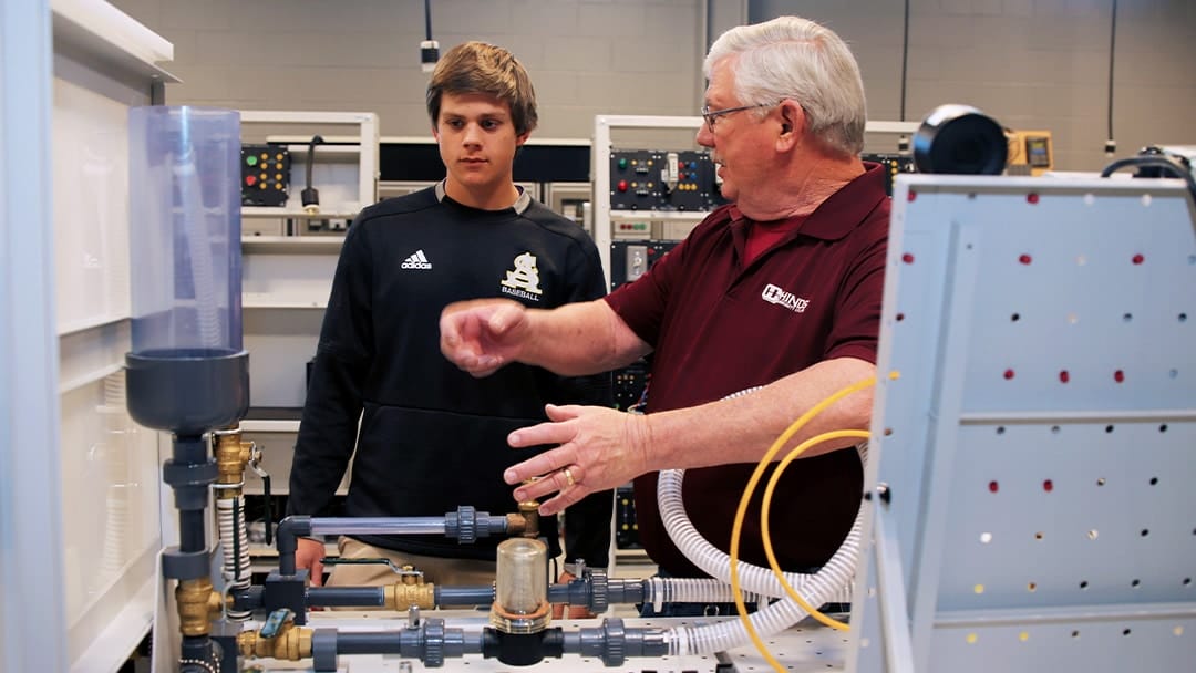 A college faculty member with grey hair explains how an industrial maintenance training apparatus works to a male high school student