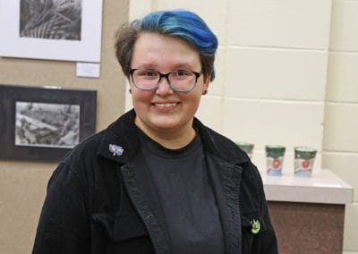 Young female college student with short blue hair standing against art gallery wall with photos and ceramics in the background