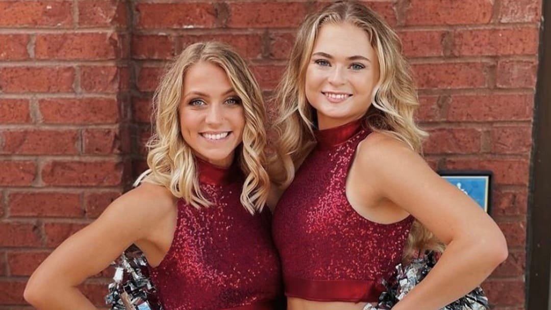 Two young female college students dressed in athletic dancing uniforms against a red brick wall