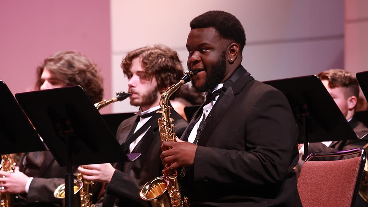 A young black male dressed in a tuxedo playing the saxophone at a band concert